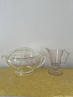 Two clear glass items side by side on a yellow surface: a covered casserole dish with queen anne division design and a small syrup/creamer pitcher