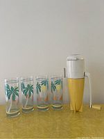 Photo showing one white and yellow Dazey Rocket ice crusher alongside four tall clear glasses with tropical prints depicting palm trees and fruits.