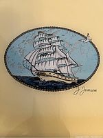 Close-up of oval foil art depicting a white sailing ship on blue sea with a bird and signature 'J Jensen'