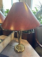 Photo of a table lamp with a conical brown leather lampshade and polished brass base seen from the side on a reflective surface near chairs and plants.