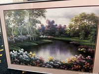 Landscape painting with a calm pond, birch trees, green grass, and colorful flowers in the foreground, custom framed with light beige mat and glass.
