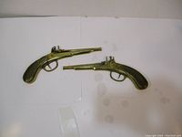 Two brass wall hanging pistols laid side by side on white surface, showing overall length and condition.