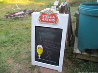 Front view of the double-sided sandwich chalkboard sign outdoors on grass, showing chalk writing and Stella Artois logo panel on top and beer glass graphic.