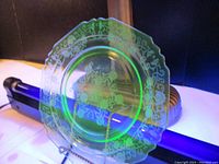 Photo of uranium glass plate front showing etched floral and scrollwork design under UV light for fluorescence effect.