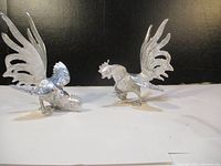 Two silver-tone metal rooster figurines positioned as if fighting, shown against a black background with white surface.