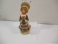 Front view of ceramic pin cushion and thimble holder figurine featuring a woman seated with pins in her hat shaped cushion.