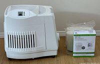 Front view of the white Aircare MA1201 evaporative humidifier displayed on hardwood floor beside three new filter packs in original packaging.