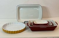 Set of all ceramic bakeware including four nested red Lagostina dishes, large white Dansk baking dish, and yellow Emile Henry quiche plate arranged side by side.