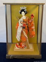 Geisha doll in display case with clear view of front, highlighting orange kimono and wooden base