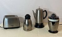 All four items: Master Chef two-slice toaster, Thermos carafe, Cuisinart coffee percolator and citrus juicer arranged side by side on a light-colored cloth.