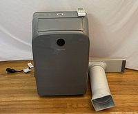 Gray portable Hisense air conditioner connected to a white flexible exhaust duct and a window vent panel, placed on wooden floor.