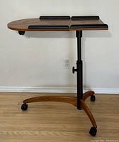 Side view of overbed table showing teak finish wood surface with leather bumpers, metal adjustable height support, and wheeled wooden base.