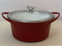 Front and side view of red cast aluminum Corningware pot with glass lid, showing ribbed exterior and metal knob handle on lid.