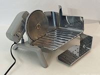 Front view of the electric slicer showing stainless steel blade, tray, and gray base with power cord.