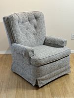 Front angled view of grey tweed upholstered swivel rocker armchair showing fabric texture, button tufting and skirted base.