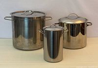 Three stainless steel stock pots of varying sizes with lids, arranged side by side showing relative sizes and handles.