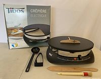 Electric crepe maker with wooden and plastic utensils laid out along with original box showing the product and brand.