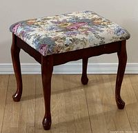 Front angle showing wooden stool with floral cushioned seat and curved wooden legs on hardwood floor.