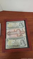 Framed set of three Canadian banknotes: two 1 dollar bills and one 2 dollar bill stacked vertically inside a wood frame.