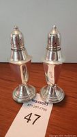 Pair of sterling silver weighted salt and pepper shakers shown from front angle on wooden surface with MaxSold auction label in view.