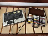 Carrying case open with 20 assorted cassette tapes inside on a wooden surface next to the cassette player.