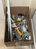 Cardboard box filled with assorted tools and hardware components including a small yellow-handled hacksaw, metal frames, garden trowel, and multiple small parts and hardware items.