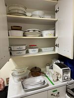 Full view of a kitchen cabinet interior showing multiple shelves with stacked white ceramic baking dishes, some with floral patterns and glass lids, including pie dishes and casserole dishes in assorted sizes.