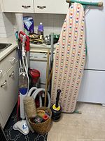 Wide view of lot showing vacuum cleaners, ironing board, basket with cleaning tools, and other supplies in kitchen area.