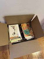 Top-down view of open cardboard box filled with a large quantity of assorted greeting cards. Cards are stacked in two piles, some in envelopes and some loose.