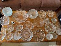 Full overview of 22 assorted clear glass trays and plates of various shapes and sizes arranged on a wooden surface