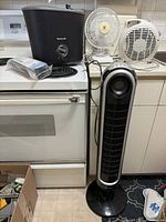 Image showing Honeywell black humidifier, small Westinghouse fan, white circular Lasko fan heater, and Honeywell tower fan in a kitchen setting on stove and counter.