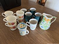Lot of nine ceramic mugs and one ceramic pitcher on a wood table, various floral patterns and colors visible.