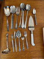 Photo showing mixed collection of 14 serving utensils including spoons, forks, a ladle, pie server, and slotted serving spoons arranged on a wooden surface.