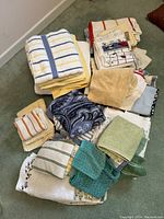 Full view of the mixed lot of various towels in different colors and patterns stacked on green carpet.