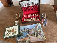Photo showing the silverware box with various utensils arranged inside, two placemats with village and bird motifs, small woven basket, two sets of coasters, and a small framed print.