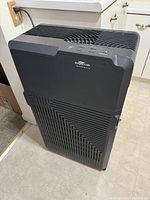 Front view of black RespirAide air purifier fan plugged in on kitchen floor next to cabinets, showing front grille and power cord.