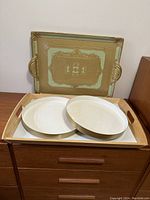 Four trays displayed on a wooden dresser against a wall: a square decorative metal tray with green and gold ornate design and handles, a rectangular wooden tray with handles, and two white oval serving trays.