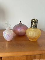 Three glass bottles on a wooden surface: a pink and white speckled perfume bottle with hummingbird stopper, a rounded iridescent pink and purple bottle with metal pump, and a yellow frosted bottle with silver decorative screw cap.