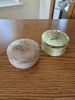 Two round stone trinket boxes side-by-side on wood surface, showing their lids with floral designs and gold trim