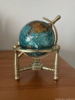 Front view of the small globe showing the colorful stone and resin continents with gold grid lines on a brass stand.