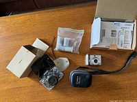 Overview of lot contents on table including Canon PowerShot A570 IS digital camera, manuals, film rolls, and camera cases