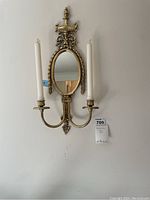 Front view of a brass wall-mounted candelabra with two candles and an oval mirror with detailed decorative frame.