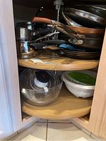 Photo showing assorted cookware including pans, bowls and a plastic colander on rotating wooden shelves inside a cabinet.