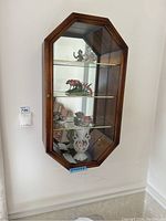 Photo showing the front view of the octagonal wall hanging curio cabinet mounted on a white wall with glass shelves inside and a glass door with a brass knob.