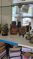 Display of the ethnic decor items on glass shelf showing the pottery vases, covered ceramic box, glass vessel, metal plate, and part of brass candle holders.