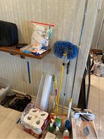 Photo of humidifier, various cleaning tools (blue dust mop, duster, mop handle), cleaning liquids, toilet paper pack, dustpan and brush, and small health items on shelf and floor near wall.
