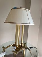 Full view of the gold toned table lamp with fabric shade, showing three cream candle-like vertical supports and a tapered cream fabric shade with gold trim.