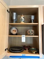Wooden cabinet shelf with 2 pewter cups, 1 ceramic stein with lid, 2 decorative plates on stands, stack of pewter plates, and 2 decorative bowls.