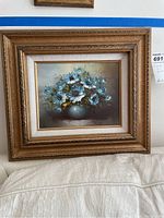 Framed oil painting showing blue flowers in a vase on a couch.