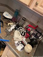 Photo showing multiple kitchen appliances and accessories on a countertop including sandwich press, hand mixer, food processor, mandoline slicer, and coffee dripper.